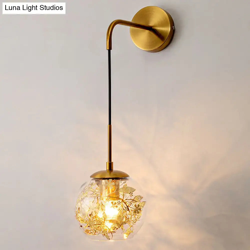Contemporary brass wall lamp with clear glass globe and gold foil flower decor featuring an illuminated filament bulb