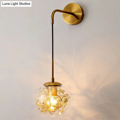 Contemporary brass wall lamp with clear glass globe and gold foil flower decor featuring an illuminated filament bulb