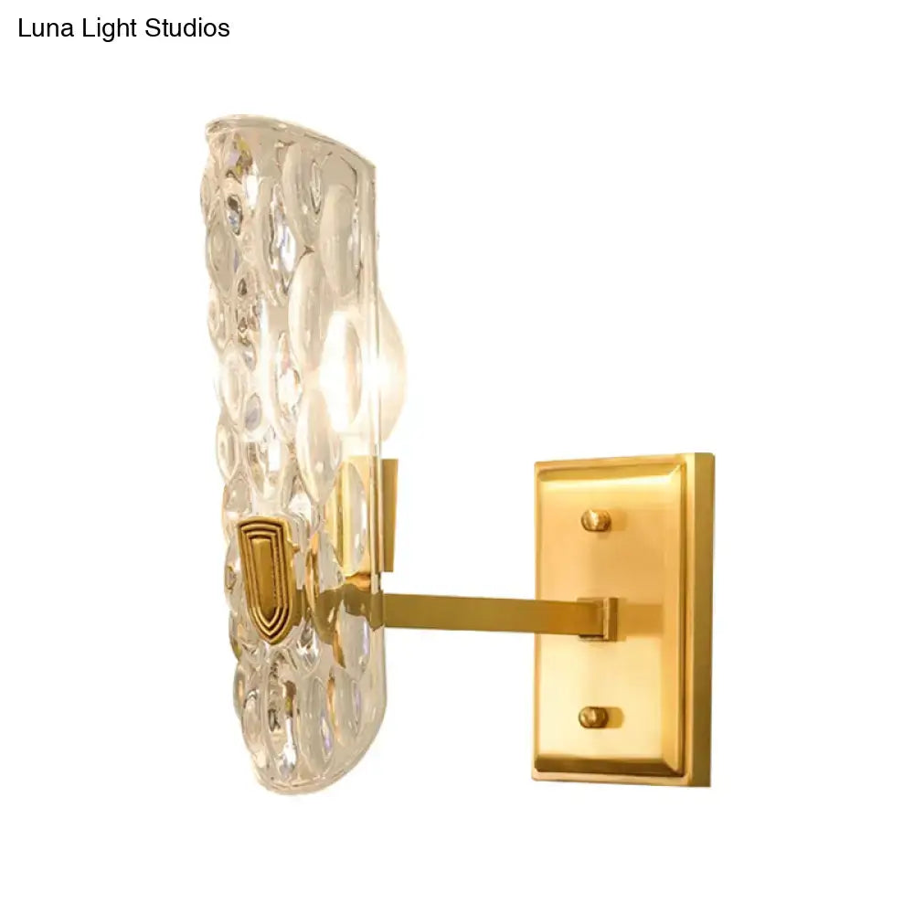 A sleek gold-toned contemporary brass wall sconce with a clear water glass shade and embossed shield detail