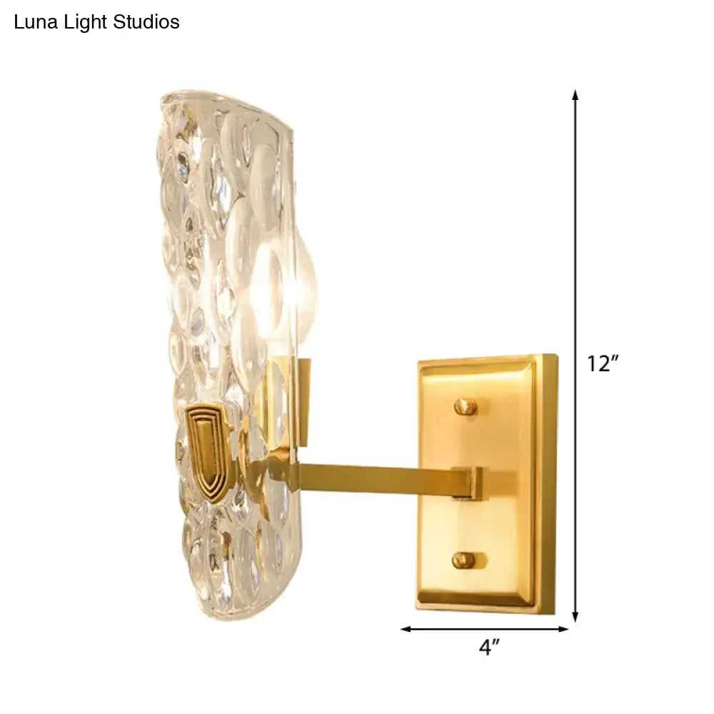 Contemporary brass gold wall sconce with clear water glass shade featuring textured embossed shield design for bedroom