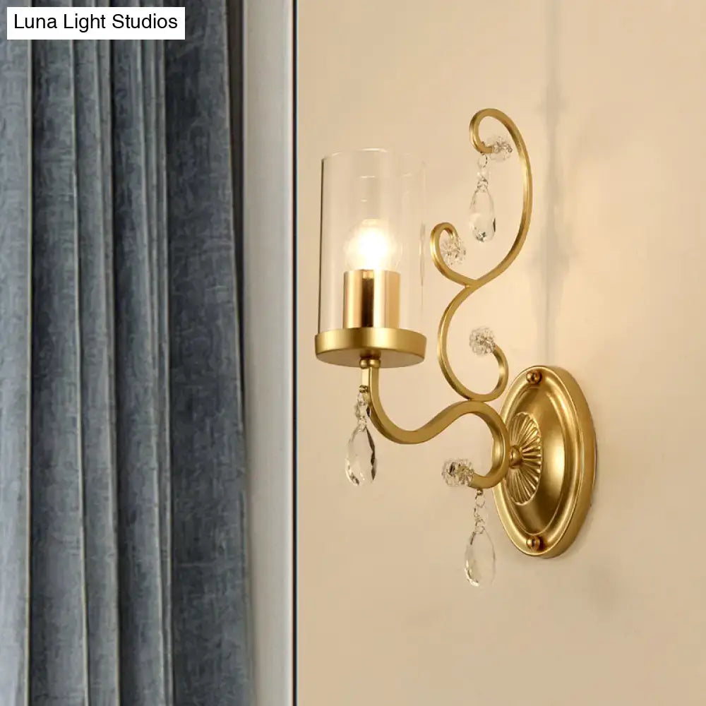 Contemporary gold-toned wall light with clear glass cylindrical shade and crystal teardrop accents for bedroom use