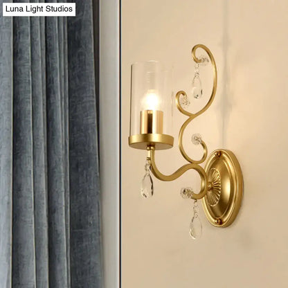 Contemporary gold-toned wall light with clear glass cylindrical shade and crystal teardrop accents for bedroom use