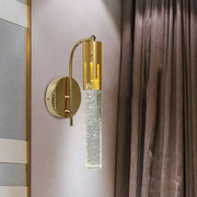 Contemporary gold-toned wall sconce with cylindrical crystal glass shade filled with sparkling particles led light bulb included
