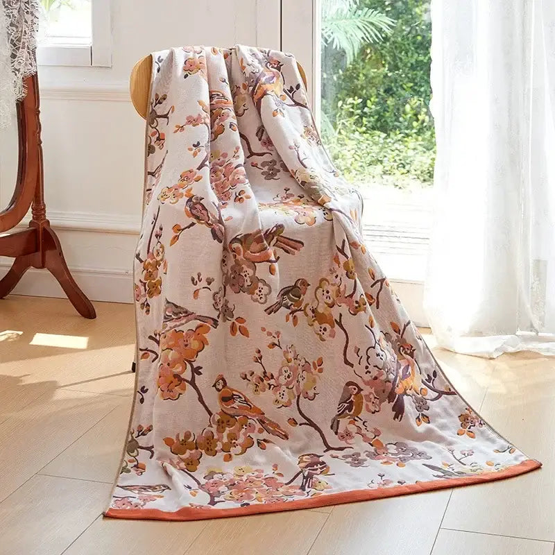 A cream-colored decorbites cotton gauze peacock bath towel with orange bird pattern draped over a wooden chair