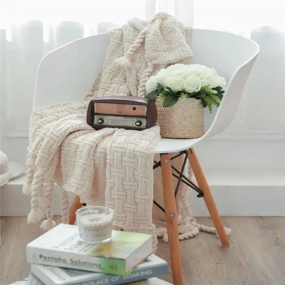 Cream-colored cozy fringed plaid knit throw blanket with textured weave and tassels draped over white chair decorbites
