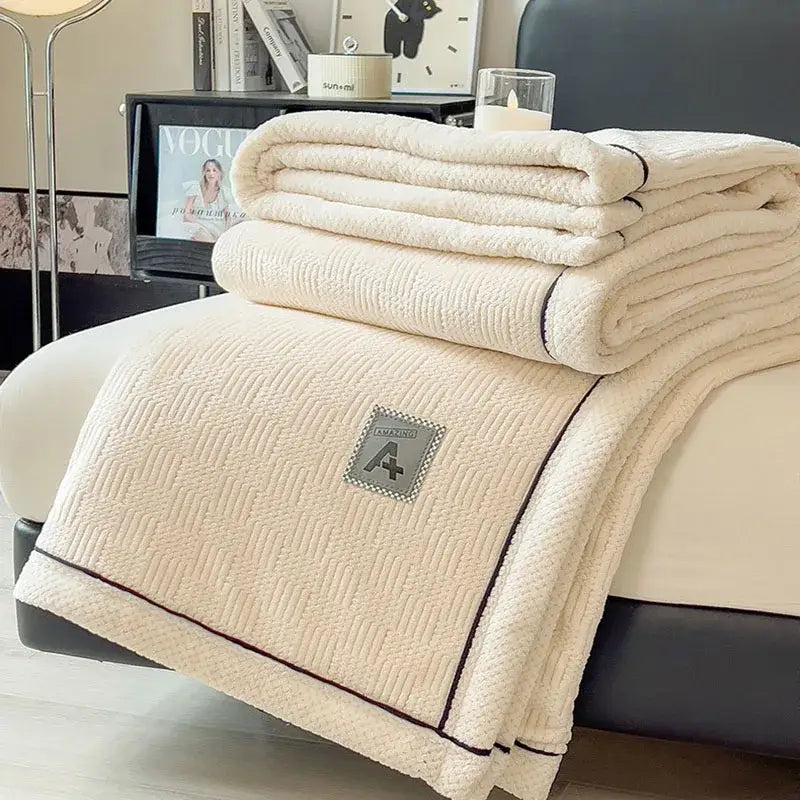Cream-colored cozy jacquard plush blanket with textured quilted pattern black piping and gray a+ embroidered patch