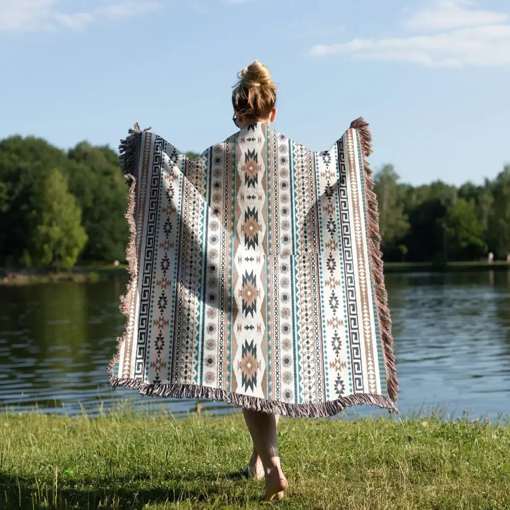 A person wearing a beige teal and black patterned decorbites cozy nordic camping blanket with fringe edges