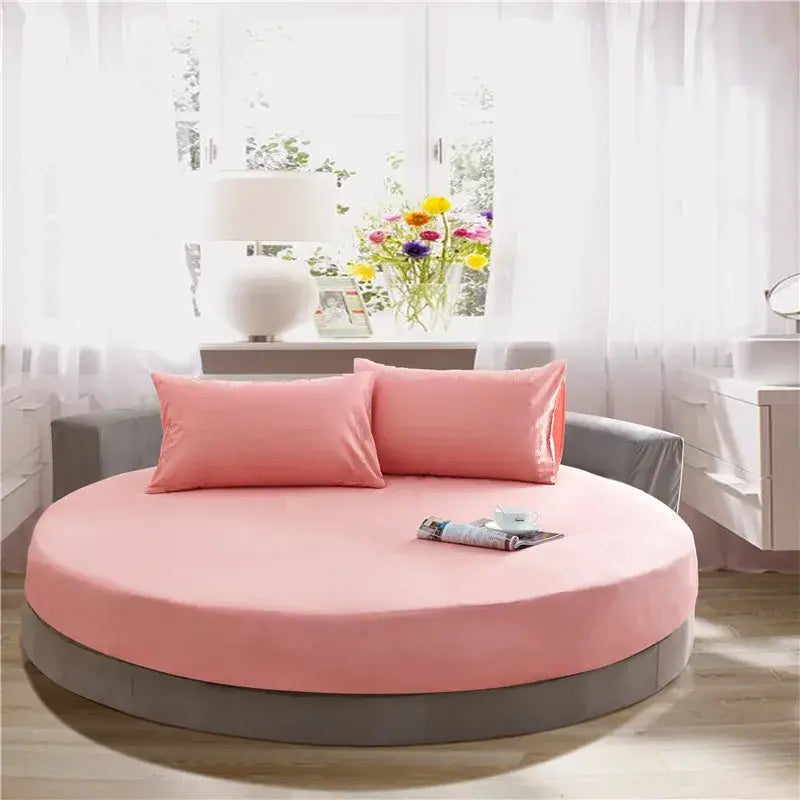Pink circular bed with gray base and matching pink pillows featuring a cream and gray round fitted bed sheet set