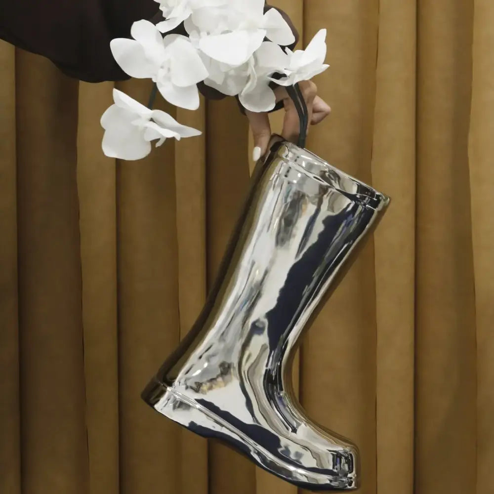 Shiny silver metallic rain boot-shaped handbag with black handle held by person against golden curtain decorbites ceramic shoe