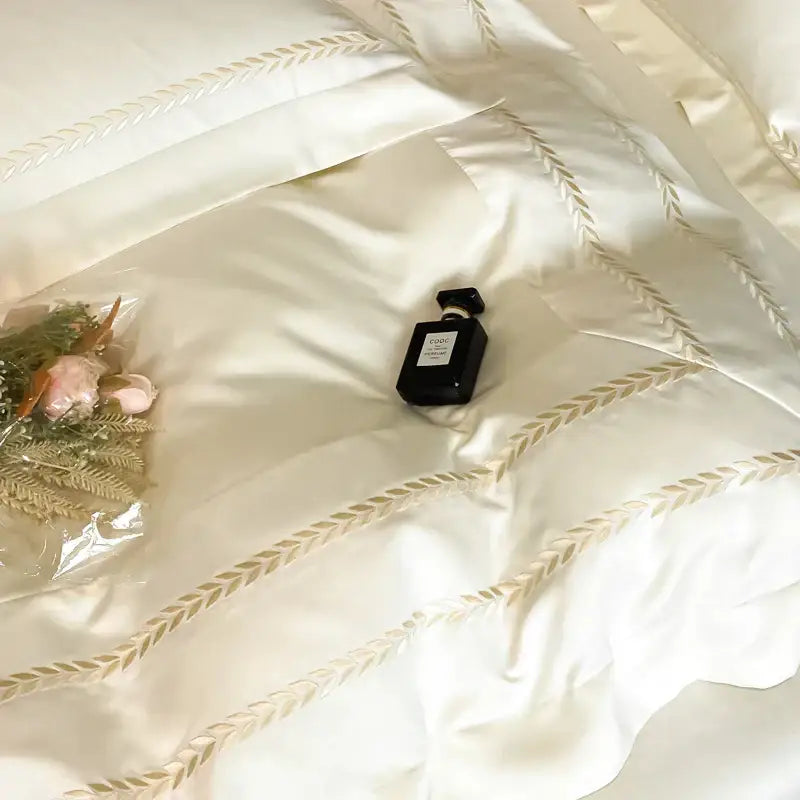 Black rectangular perfume bottle with silver cap resting on cream embroidered ice silk bedding set with gold leaf design