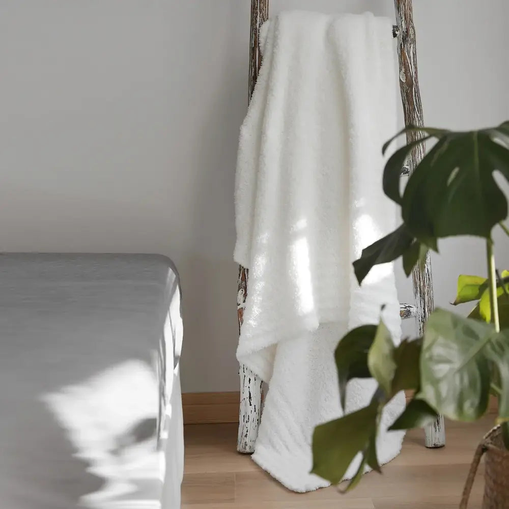 A cream knitted throw blanket made of plush microfiber draped over a rustic wooden ladder in a bedroom setting
