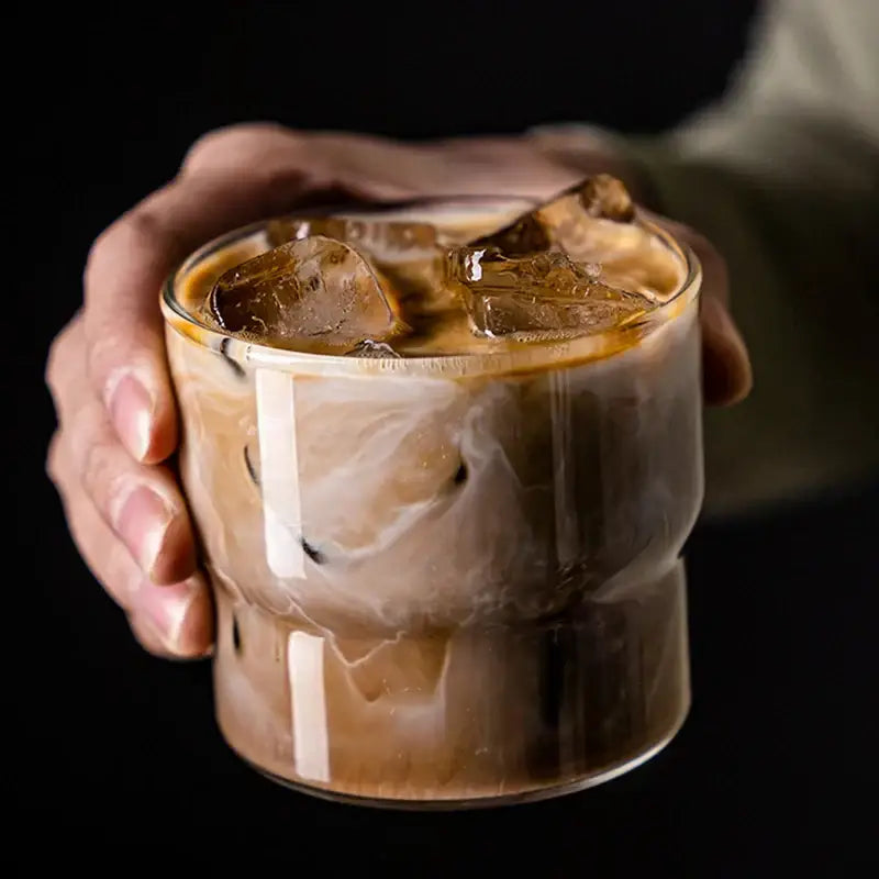 A hand holds a clear decorbites creative glass coffee smoothie cup with iced coffee swirling layers of brown and cream