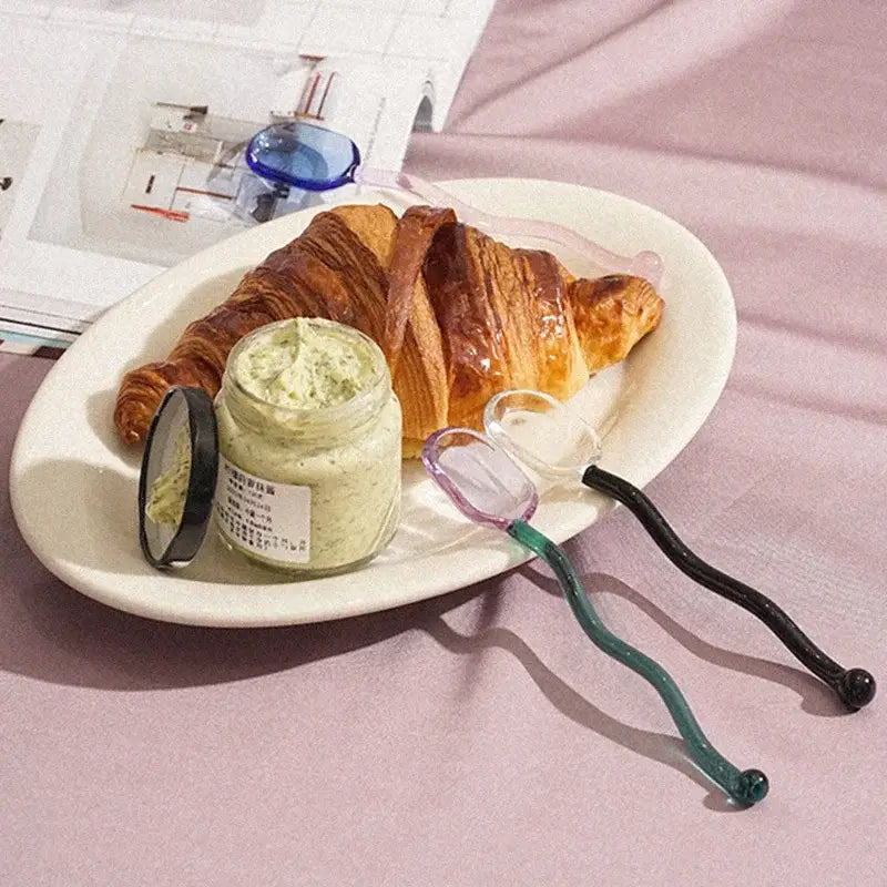 A golden brown croissant with flaky layers beside a jar of spread on an oval plate with a creative glass spoon dessert set