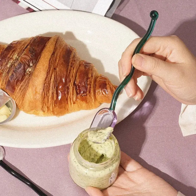 A creative glass spoon dessert set featuring a sleek dark green handle and translucent purple bowl scooping from a jar