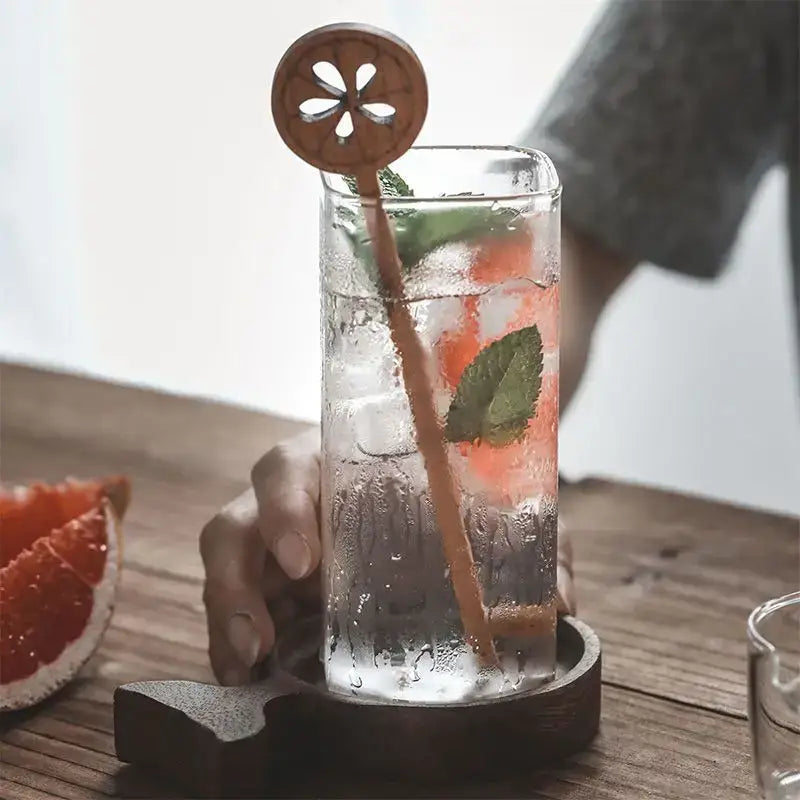A clear decorbites creative square glass with gold rim filled with pink drink ice mint and wooden citrus stirrer