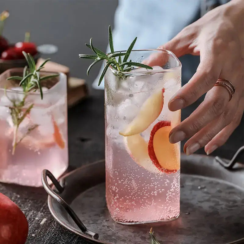 A tall clear decorbites creative square glass with gold rim filled with pink sparkling drink and ice cubes