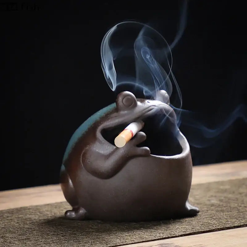 Cute big belly dark brown ceramic ashtray shaped like a frog with teal glaze holding a cigarette emitting smoke