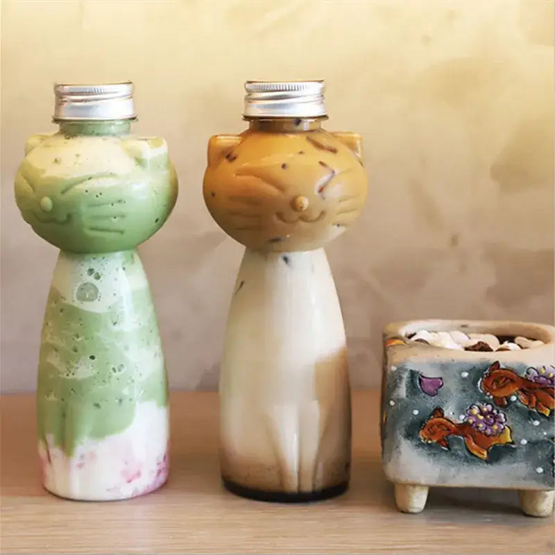 Two cute cat beverage bottles with silver screw caps containing green white and brown cream drinks for coffee and juice