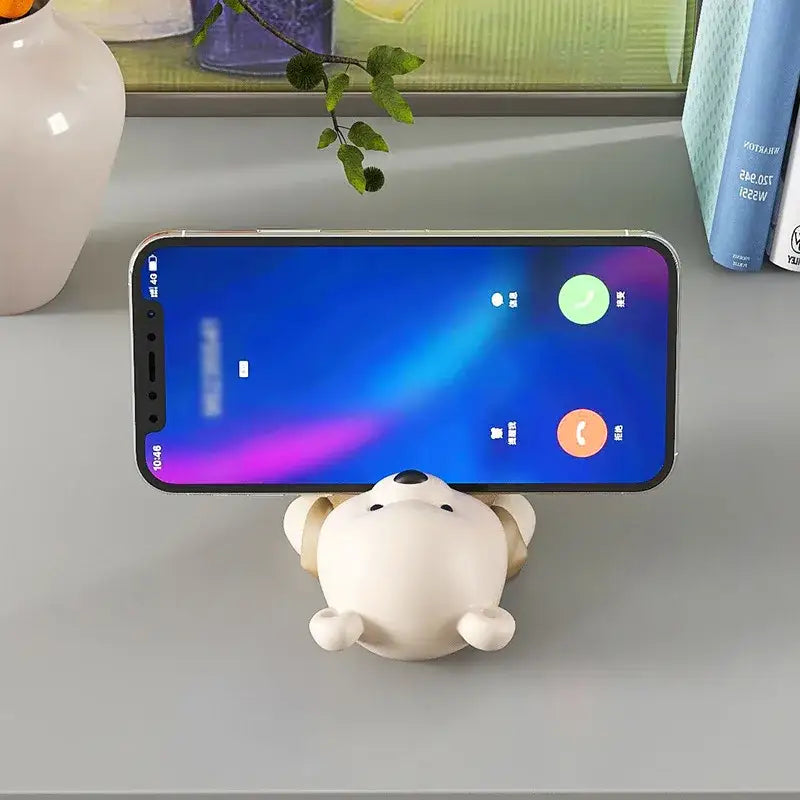 Cute cream ceramic bear cellphone stand in the shape of a white polar bear with beige shorts holding a silver smartphone upright