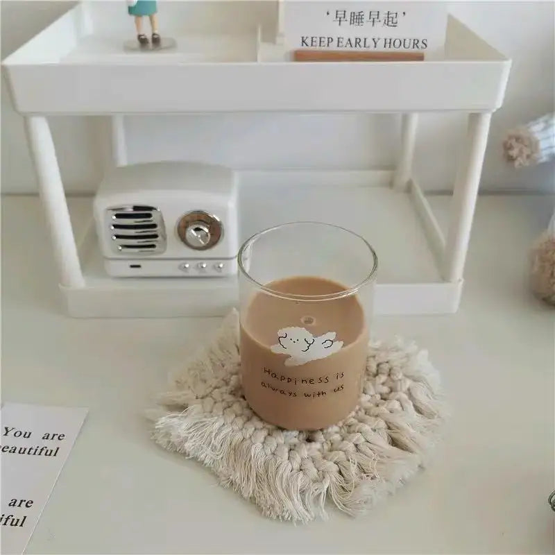 A clear glass cup filled with a light brown beverage on a cream macrame coaster featuring a cute dog glass design