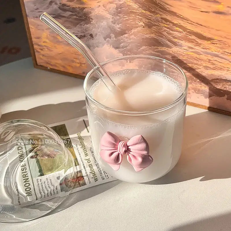 A clear glass cup with a pink bow tie filled with a creamy beverage and a matching transparent straw