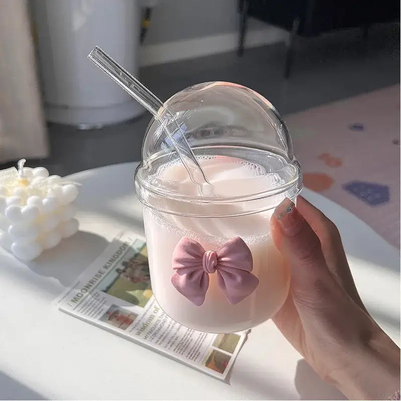 A clear glass cup with a dome lid and a pink bow tie holding a light-colored beverage for kitchen use