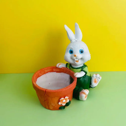 White plastic bunny rabbit figurine in green overalls sitting beside an orange ceramic rabbit flower pot with floral decoration
