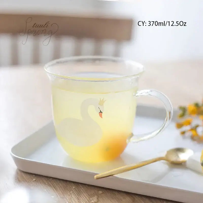 Clear glass coffee mug with white swan design and gold crown filled with yellow liquid for kitchen use