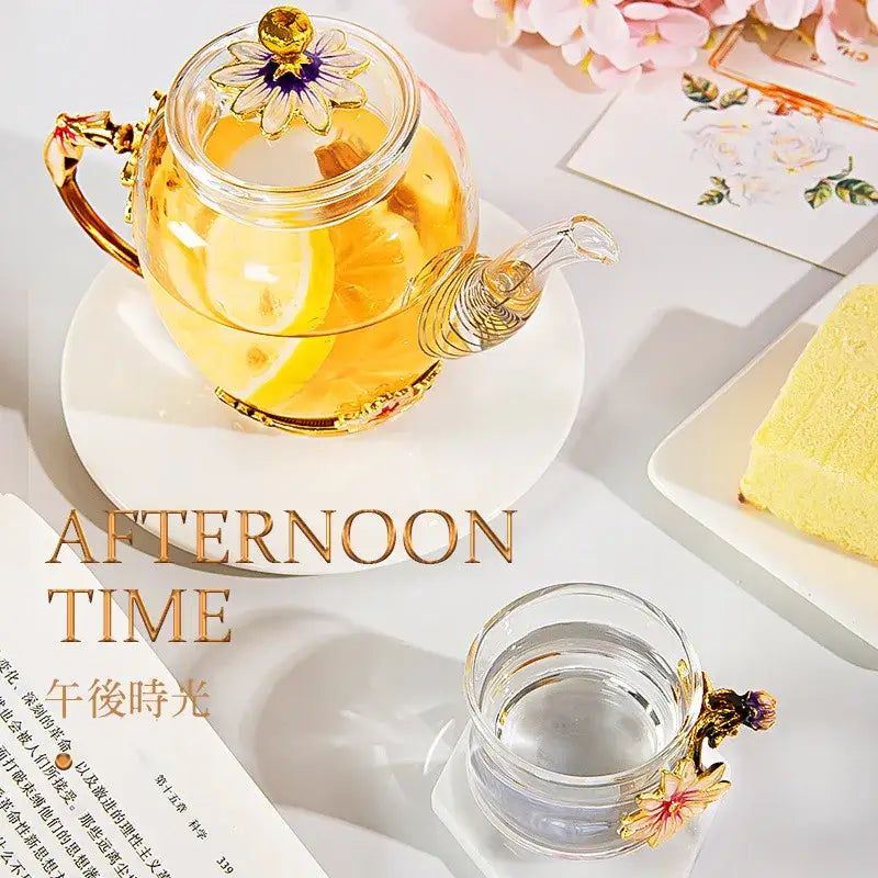 A clear glass teapot set with gold-toned handles and flower-shaped lid filled with lemon tea from Daisy enamel pink crystal glass
