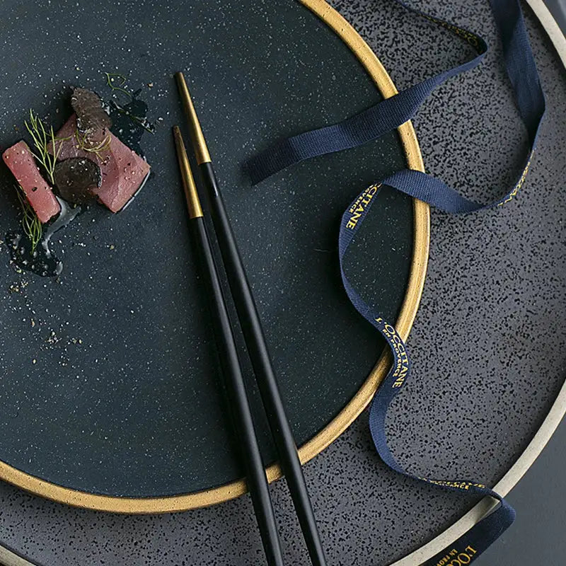 Black chopsticks with gold tips rest on a dark teal gold rimmed ceramic western dinnerware set plate by decorbites