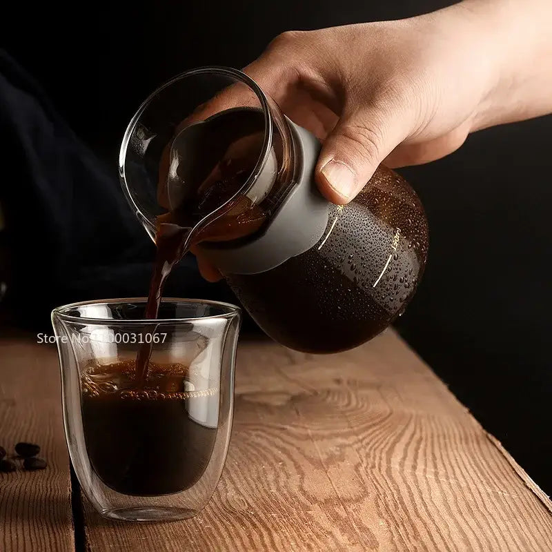 A sleek transparent glass coffee carafe with matte gray silicone handle pouring into a double wall glass coffee mug