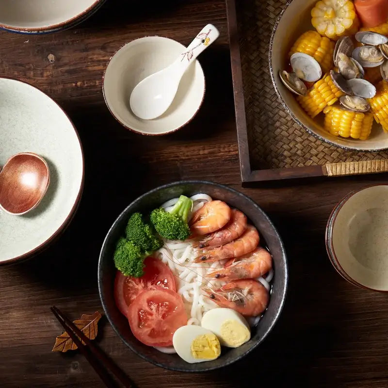 Dark gray ceramic ramen bowl filled with udon noodles topped with shrimp, tomatoes, broccoli, and hard-boiled egg