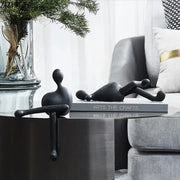 Two black minimalist abstract characters resin figurines by decorbites rest on books on a modern metallic table