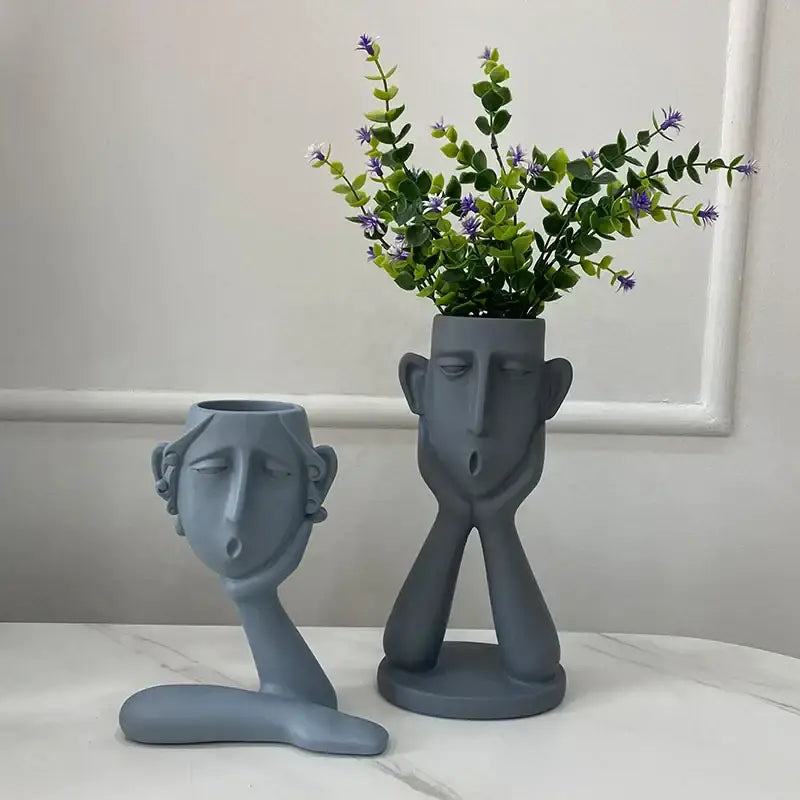 Gray ceramic face-shaped flowerpot ornament with matte finish holding greenery and purple flowers by decorbites abstract family