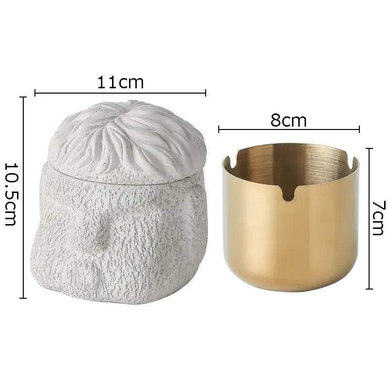 White textured skull-shaped figure portraits cement ashtray with matching lid and gold-toned metal ashtray beside it