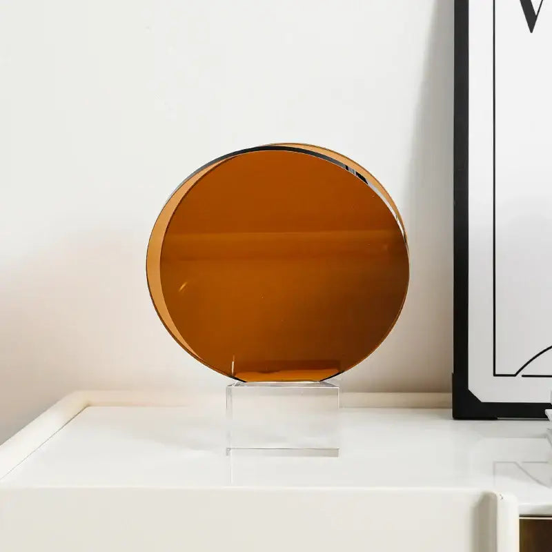 A sleek amber-tinted circular ornament with a polished metallic rim on a clear acrylic stand for acrylic crystal bookshelf decor