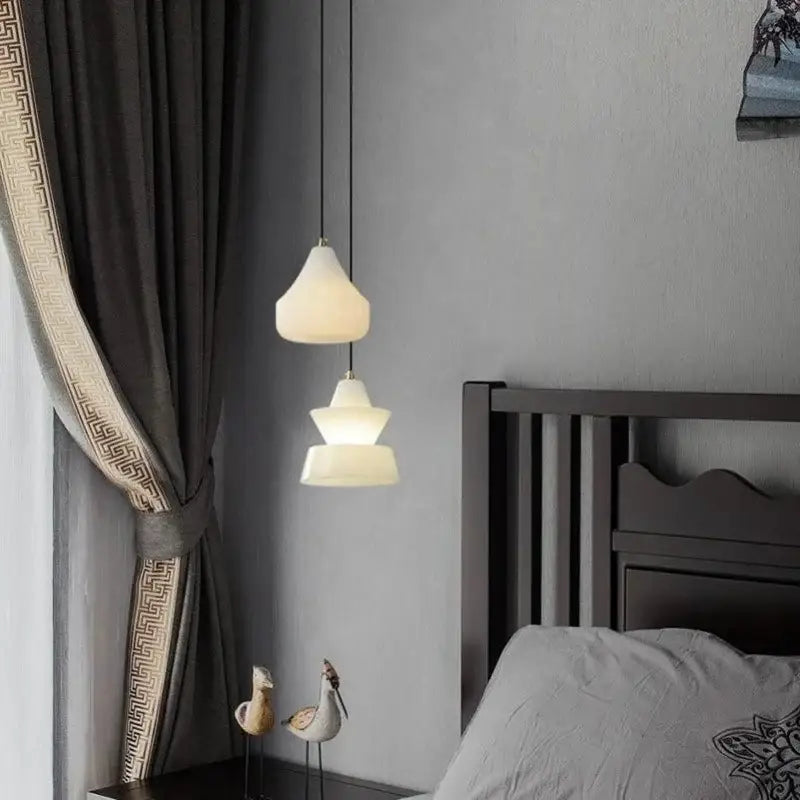 Cream-colored frosted alabaster pendant lights with a modern tiered design hang from black cords above a bed