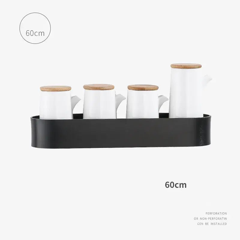 Set of white ceramic oil and vinegar dispensers with wooden lids arranged on a black tray beside an aluminum bathroom shelf with towel bar
