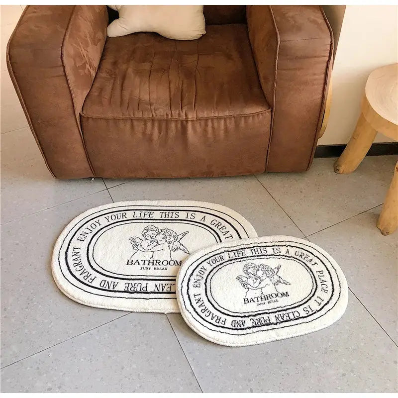 Cream-colored oval cream-colored bath rug with black embroidered angel print shaggy text and cherub illustrations