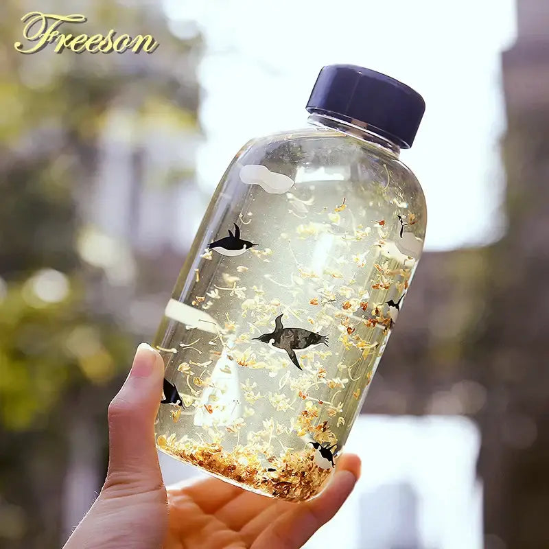 DecorBites™ glass water bottle with black penguin decals, floating dried yellow flowers, and dark blue cap for outdoor use
