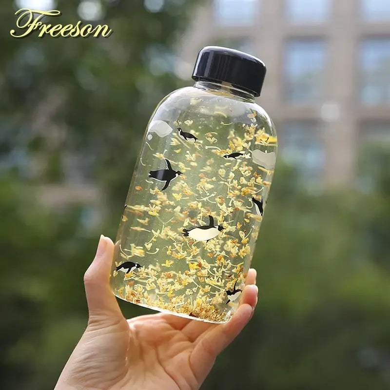 Clear glass water bottle with black lid filled with golden dried flowers and black-and-white penguin figurines from decorbites