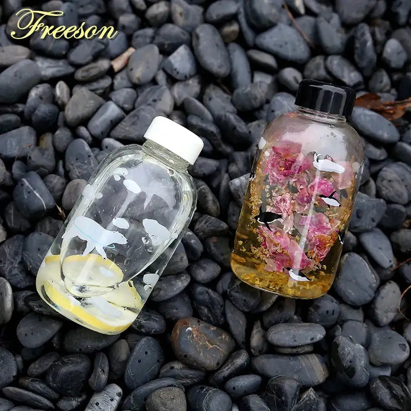 Two transparent glass water bottles with white whale silhouettes lemon slices and pink roses with black birds on dark pebbles