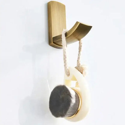 A cream oval bath brush with dark gray bristles and gold accents hanging on a brass hook in an antique brass bathroom accessory set