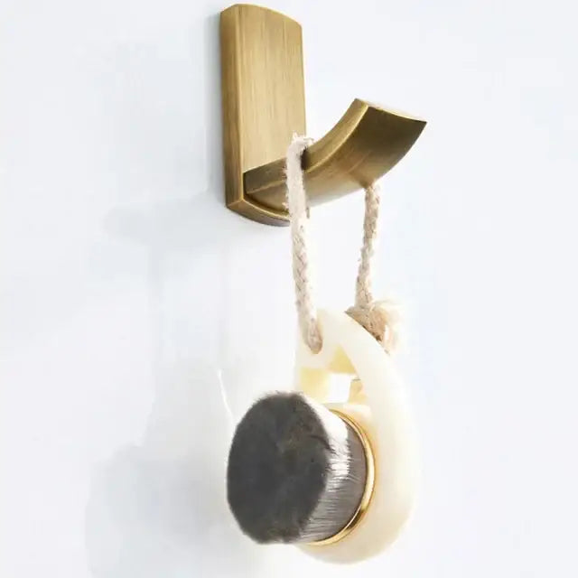 A cream oval bath brush with dark gray bristles and gold accents hanging on a brass hook in an antique brass bathroom accessory set