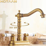 Antique brass dual basin faucet with ornate segmented spout and white ceramic handles featuring black floral detailing