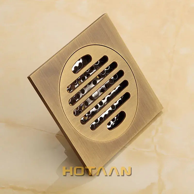 Square brass-finished floor drain with circular grille featuring horizontal slats and antique brass flower art design