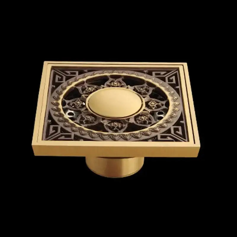 Gold-colored square floor drain in antique brass with intricate black and gold flower art filigree design for bathroom use