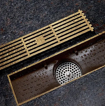 Decorbites™ antique brass linear floor drain with gold-colored rectangular metal grate and perforated circular catch basin