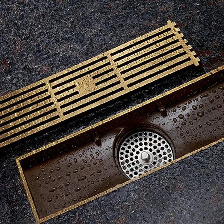 Decorbites™ antique brass linear floor drain with gold-colored rectangular metal grate and perforated circular catch basin