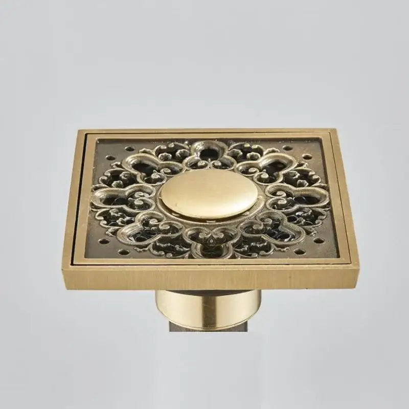 Square fish carved antique brass floor drain with ornate floral cutout and central circular button for bathroom use